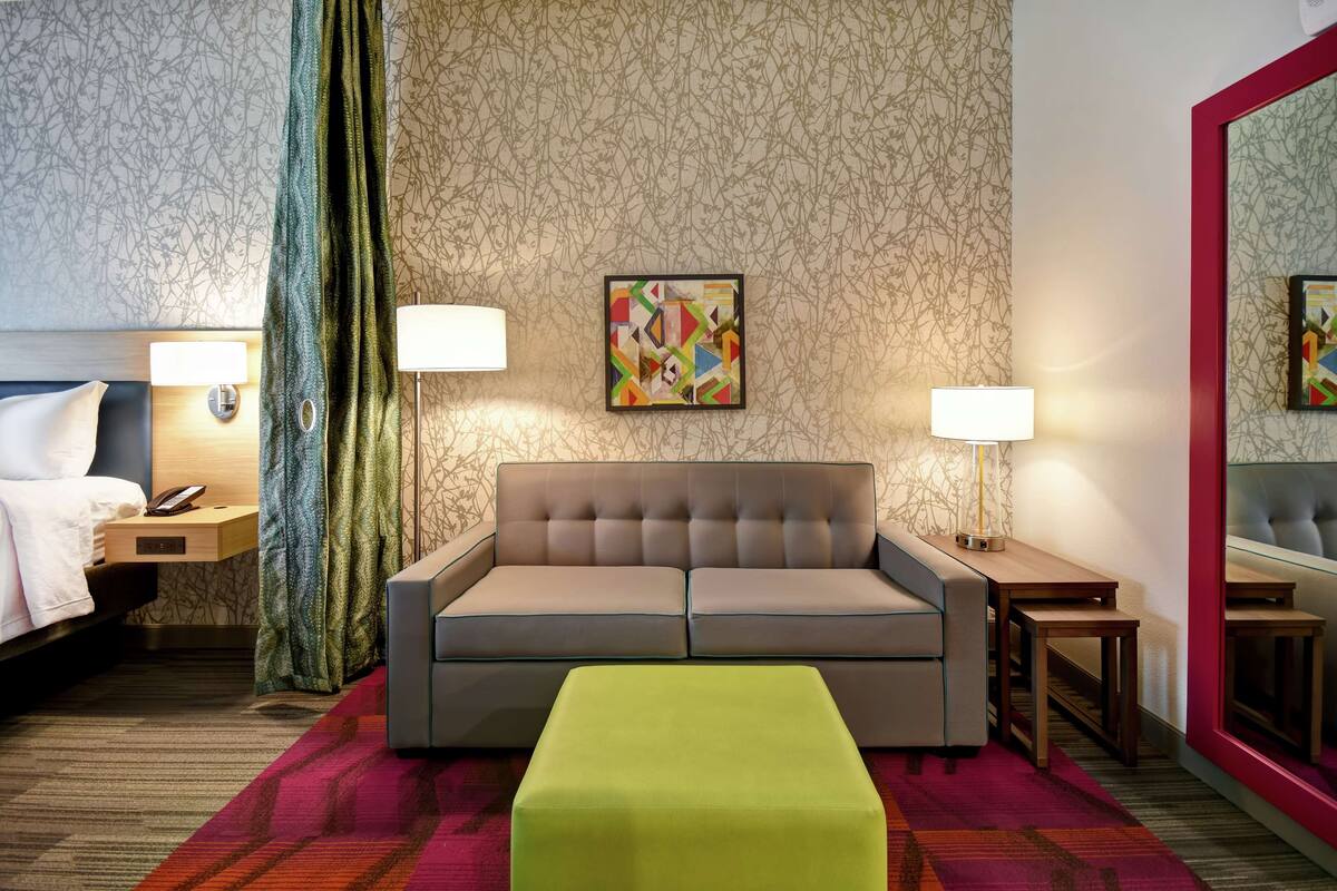 Une chambre d’hôtel avec un canapé capitonné, un pouf vert, deux tables de chevet avec des lampes, un lit recouvert d’une couette blanche et un grand miroir.