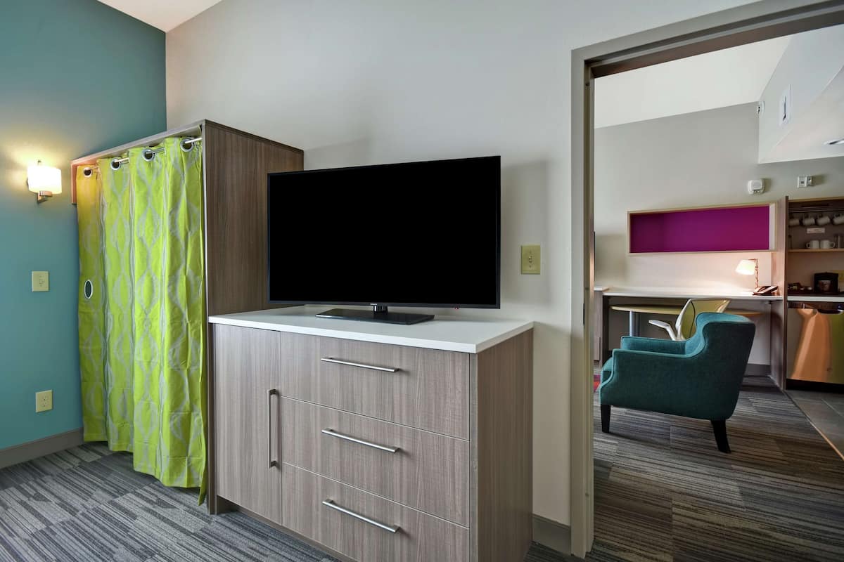 Une chambre d’hôtel avec un téléviseur à écran plat posé sur un meuble en bois, un rideau de douche à motifs verts et un fauteuil turquoise.