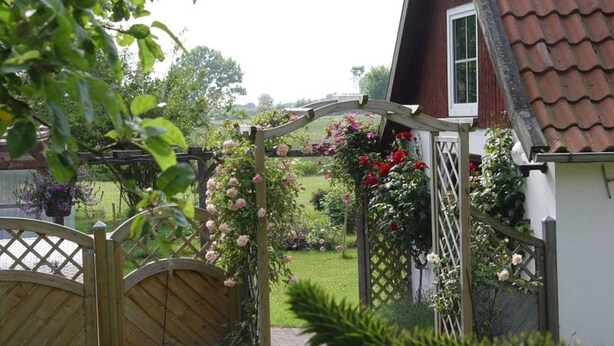 Une porte en bois mène à un jardin où se trouve un bâtiment blanc surmonté d’un toit recouvert de tuiles rouges.