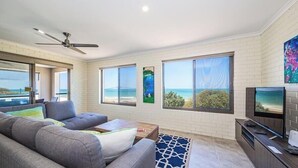 Uma sala de estar moderna com sofá, mesa de centro, TV e janelas amplas com vista para o oceano.