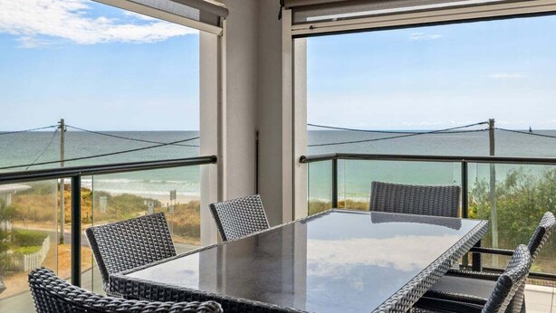 Varanda com mesa de vidro e cadeiras de vime, com vista para a praia e o oceano.