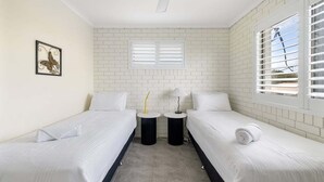 Quarto com duas camas de solteiro, ventilador de teto, janela com persianas e uma pequena mesa com abajur.