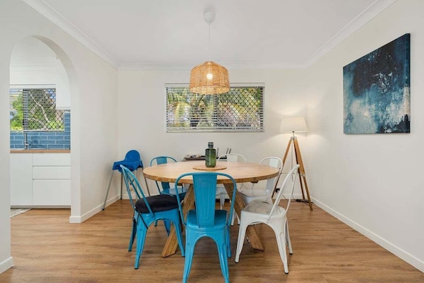 Un comedor con una mesa redonda de madera, sillas blancas y azules, una lámpara colgante de fibras, una ventana con vistas y un cuadro en la pared.