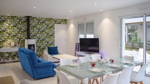 A modern living room with a blue sofa, a glass dining table set for six, and a TV in the background. - 3 bedroom awesome home in Matignon (Matignon)