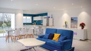 A modern living room with a blue sofa, a white coffee table, and a dining area with white chairs. - 3 bedroom awesome home in Matignon (Matignon)