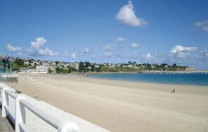 A wide sandy beach with a clear blue sky and a few clouds. There are buildings in the background and a few people walking along the shore.