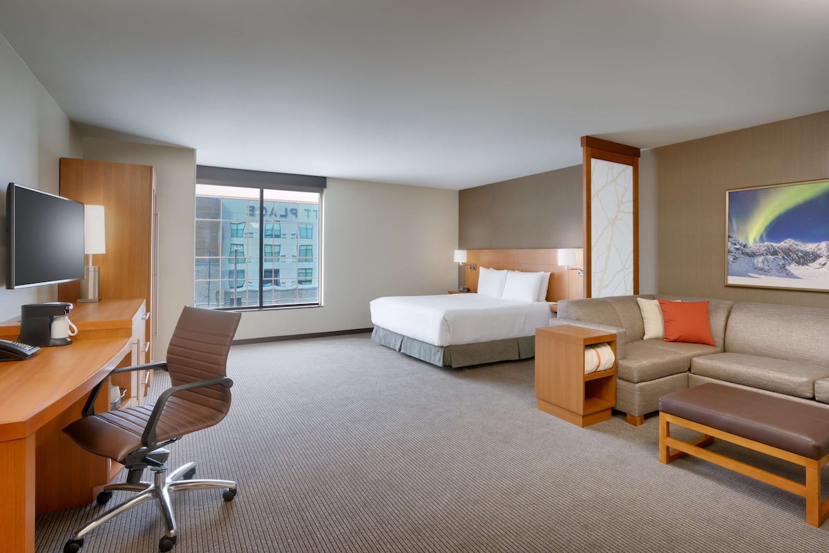 Habitación de hotel con cama, escritorio con silla, sofá, banco, televisión y ventana con vista a la ciudad.
