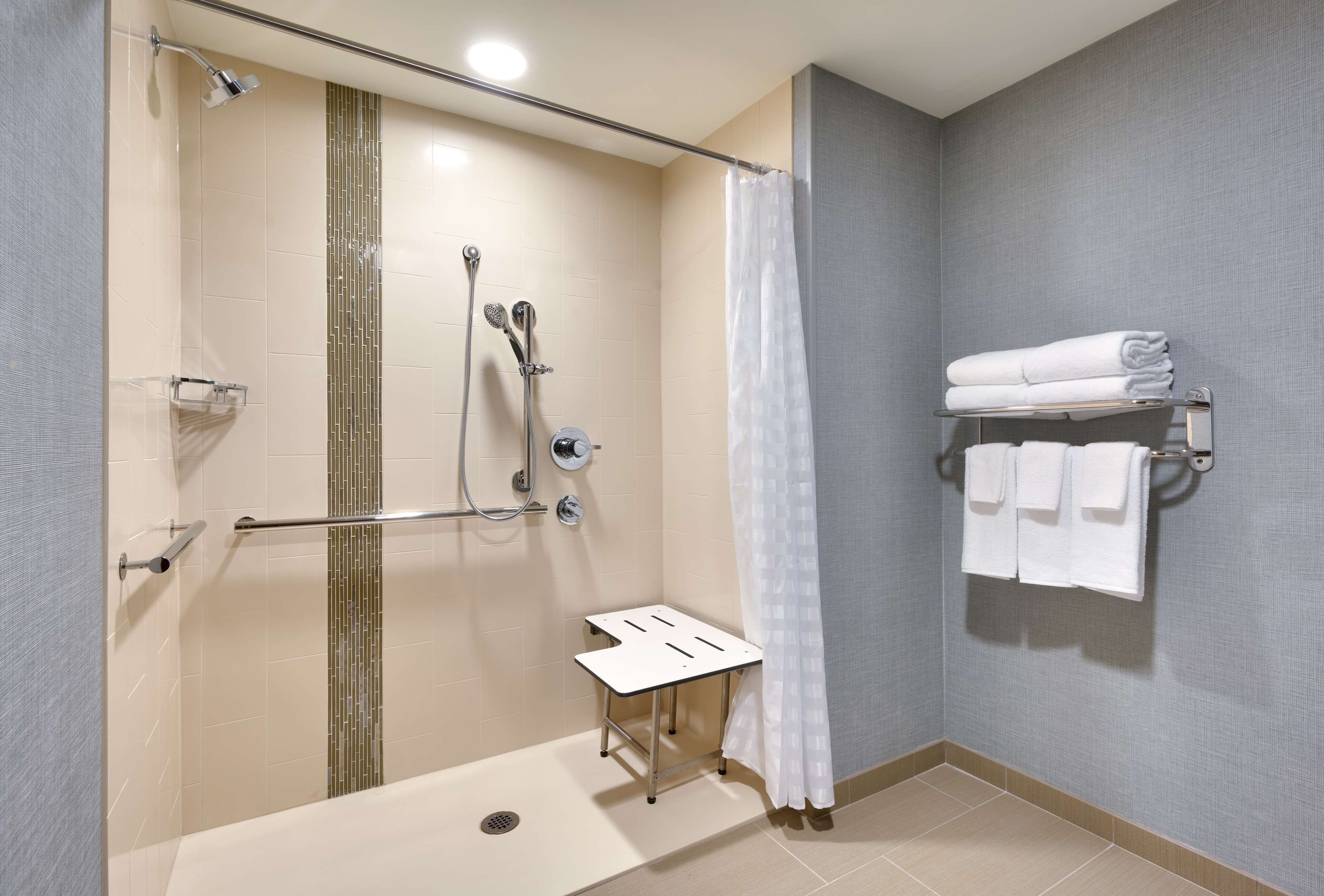 Un espace douche doté d’une porte en verre, d’un pommeau de douche fixe, d’un pommeau de douche à main, d’un porte-serviettes avec des serviettes pliées et d’une petite table.
