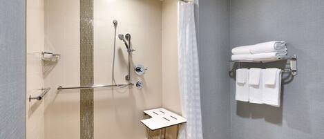 Un espace douche doté d’une porte en verre, d’un pommeau de douche fixe, d’un pommeau de douche à main, d’un porte-serviettes avec des serviettes pliées et d’une petite table.