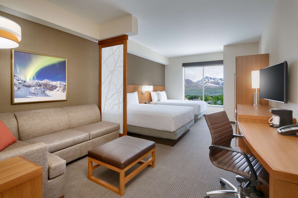 Habitación de hotel con sofá, dos camas, escritorio con silla, televisión y una ventana grande con vista a la montaña.