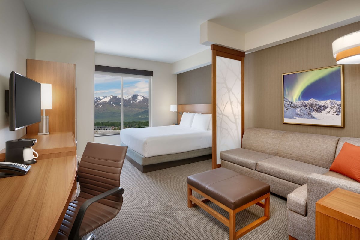 Habitación de hotel con cama, sofá, escritorio con silla, televisión y un amplio ventanal con vistas a la montaña.