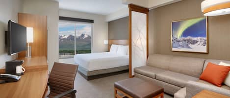 A hotel room with a bed, a sofa, a desk with a chair, a TV, and a large window with a mountain view.