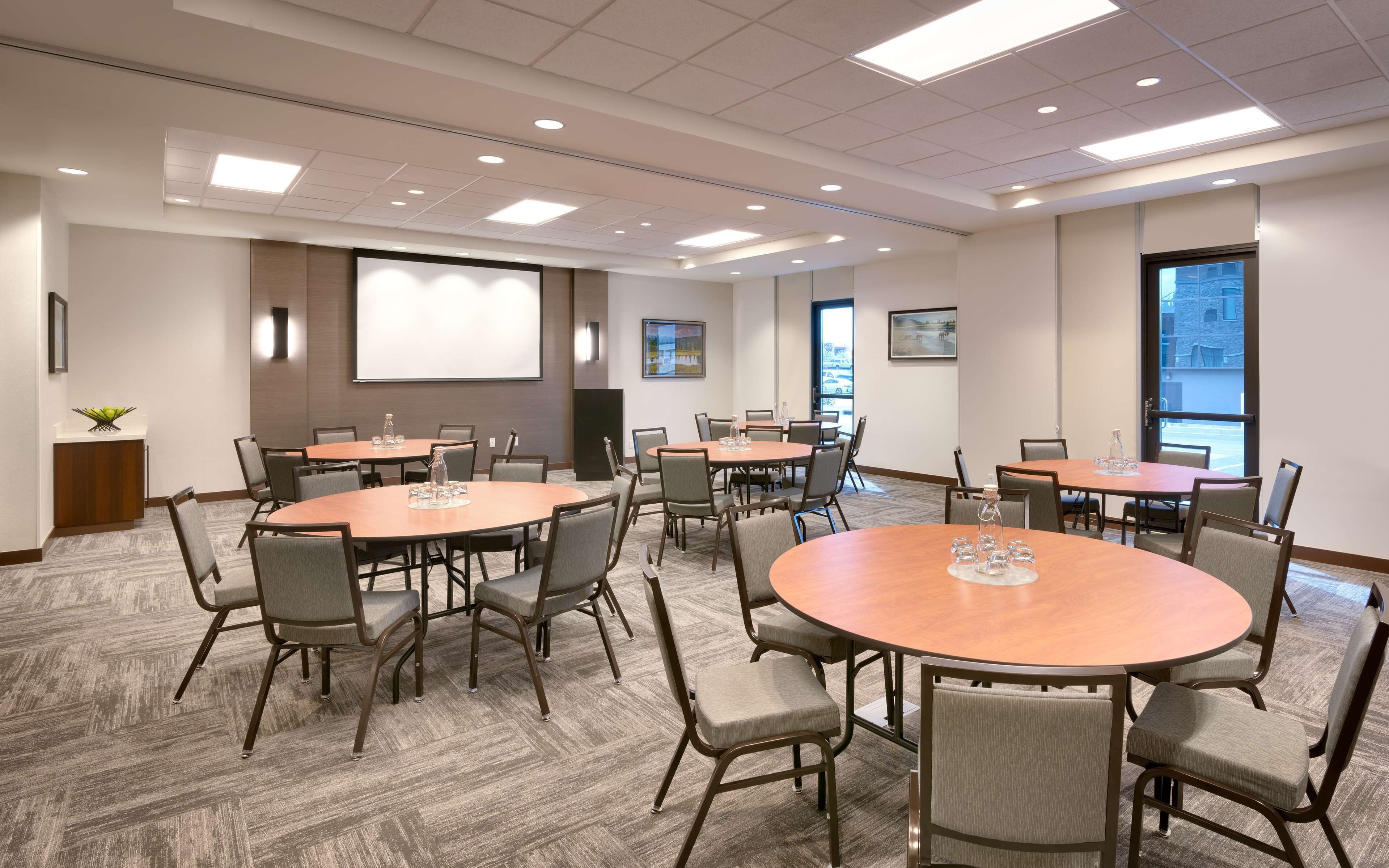 A conference room with multiple tables and chairs, a projector screen, and a large window with a view of buildings.
