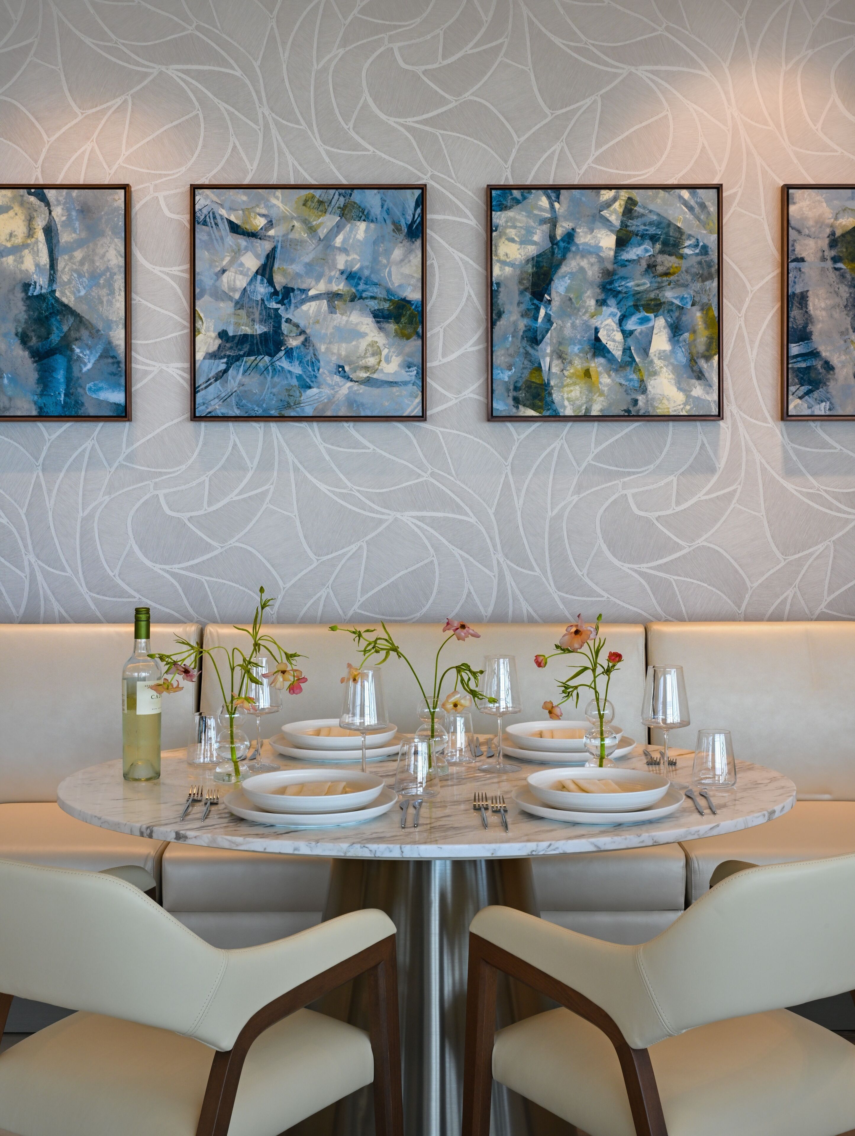A dining area with a marble table set for four, surrounded by cushioned chairs. Above the table, there are three framed abstract paintings.