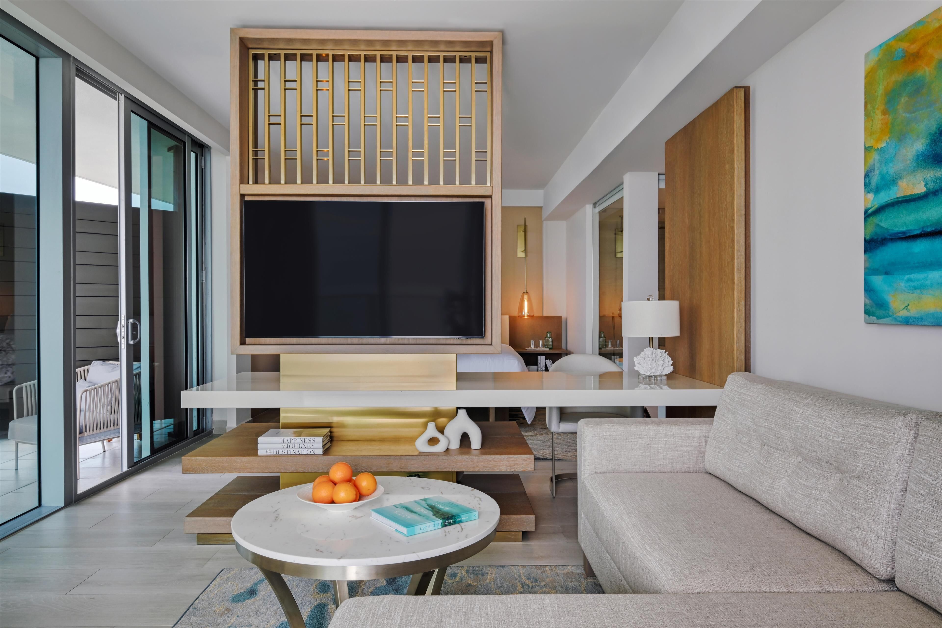 A modern living room with a flat-screen TV, a sofa, a coffee table with books and oranges, and a sliding glass door leading to a balcony.