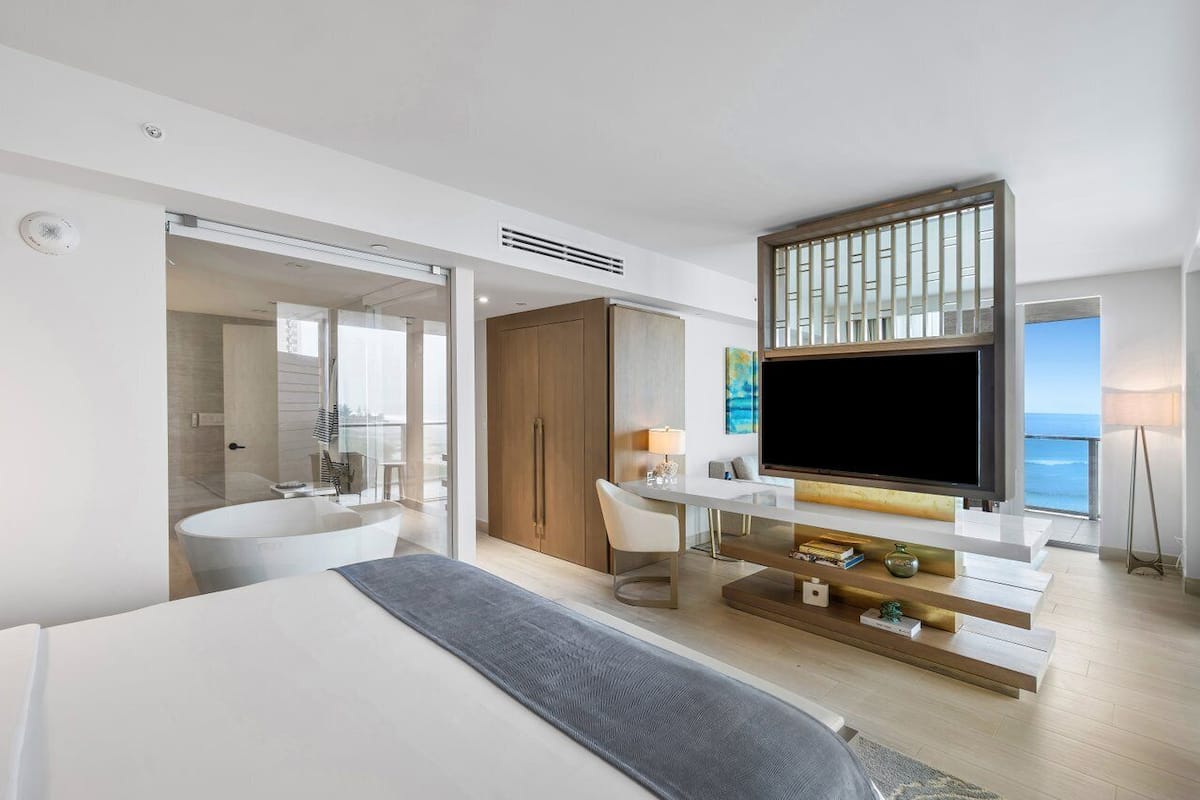 A modern hotel room with a large bed, a TV, a desk with a lamp, a view of the ocean, and a bathroom with a bathtub.