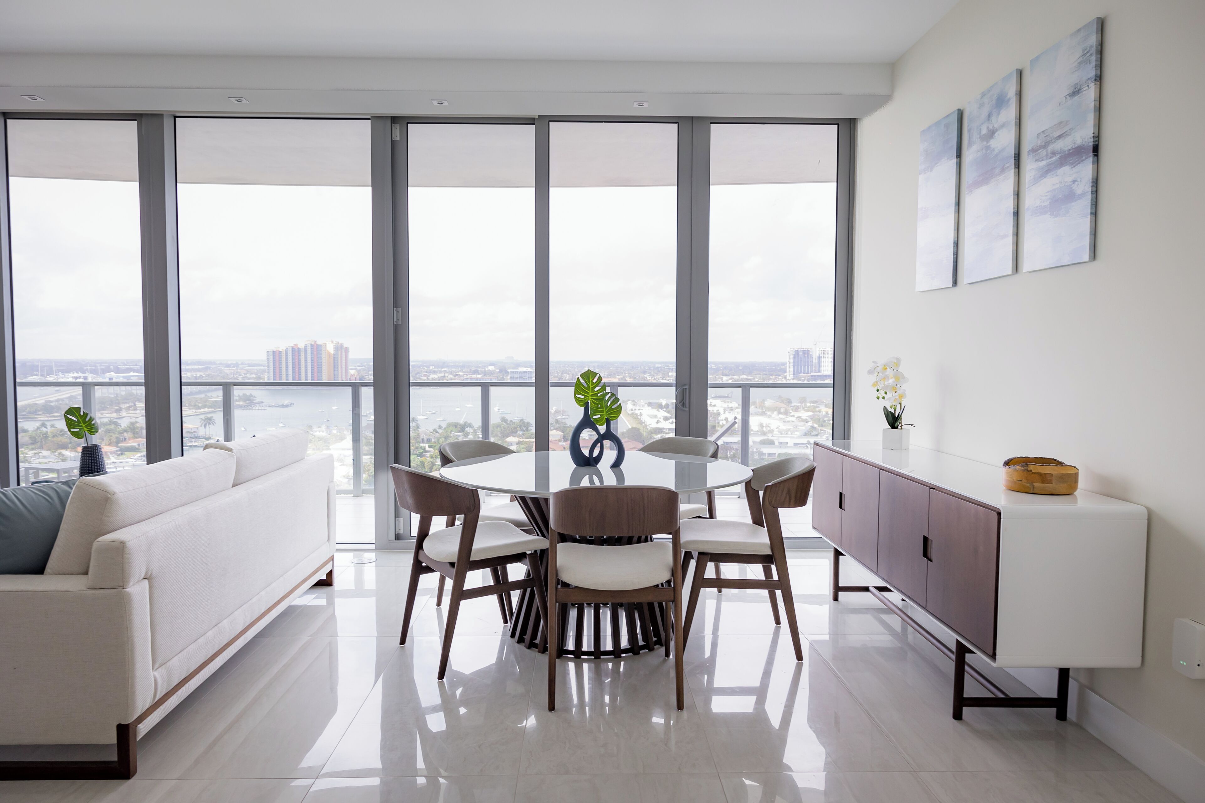 A modern living room with a dining table, white sofa, and a view of the city through large windows.