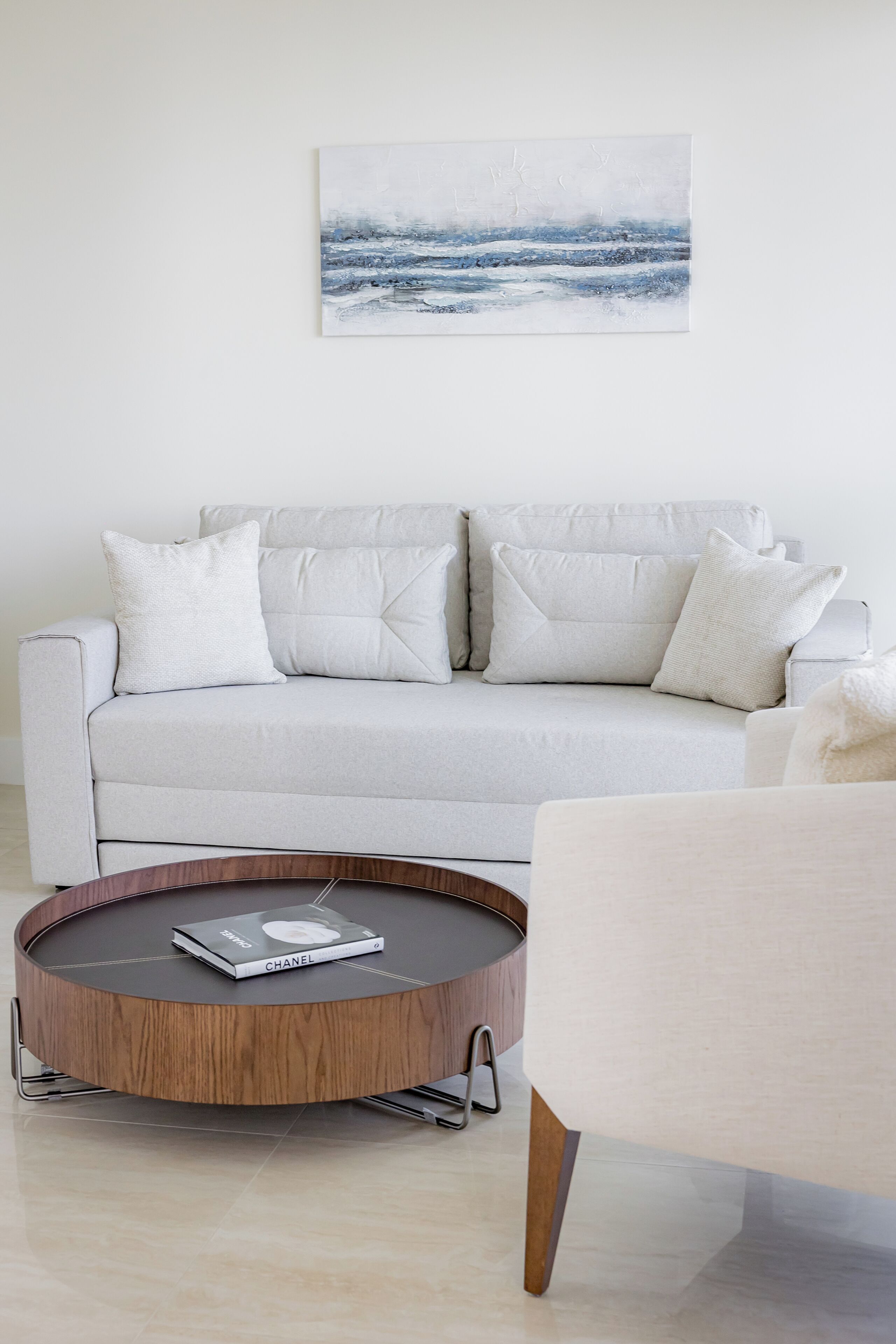A modern living room with a light grey sofa, a round wooden coffee table, and a white armchair. A book titled 'CHANEL' is on the coffee table.