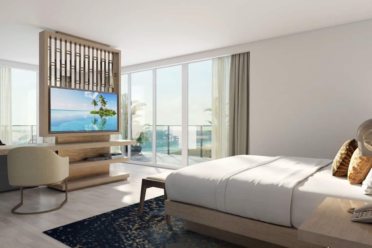 A modern hotel room with a large bed, a desk with a flat-screen TV, a balcony with a view of the sea, and a phone on the bedside table.