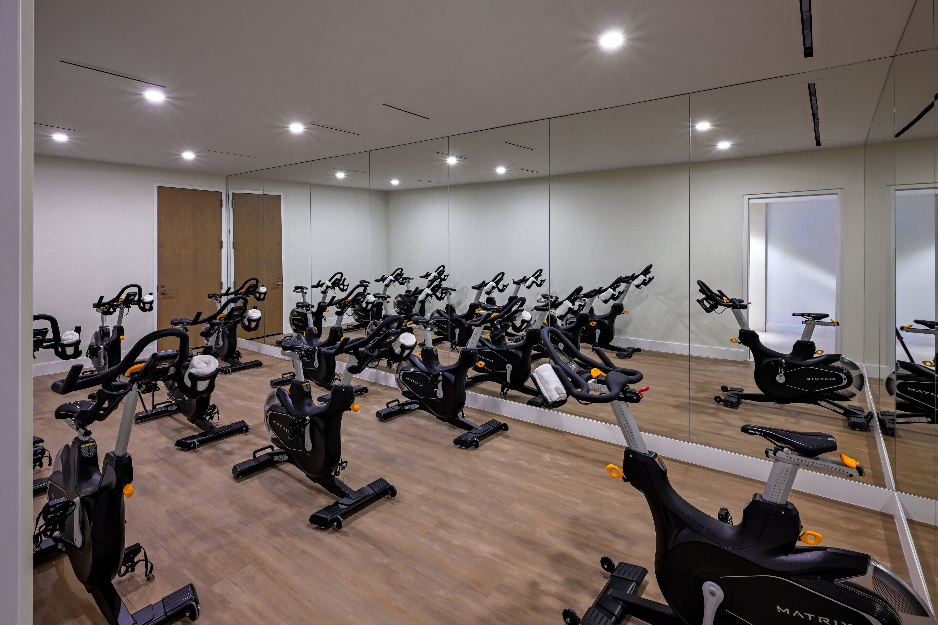 Une salle de sport équipée de plusieurs vélos d’exercice, dotée d’un plancher en bois et de murs recouverts de miroirs.
