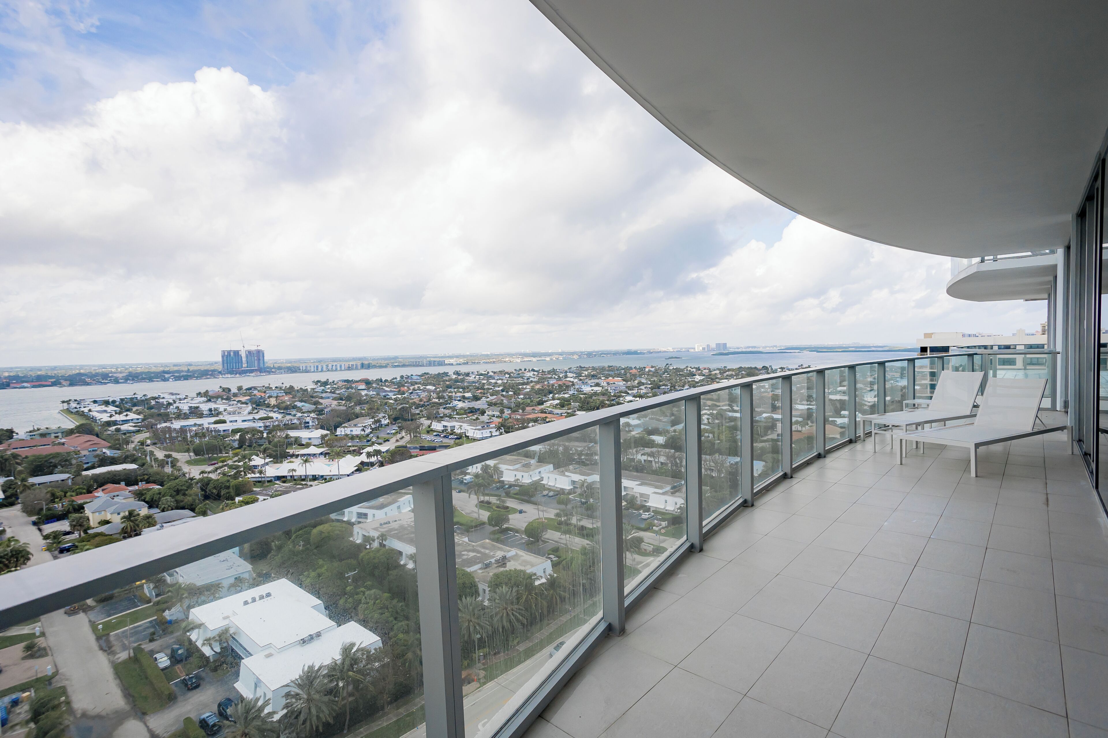 Un balcon doté d’une balustrade en verre, offrant une vue sur un paysage urbain avec des bâtiments et une étendue d’eau.