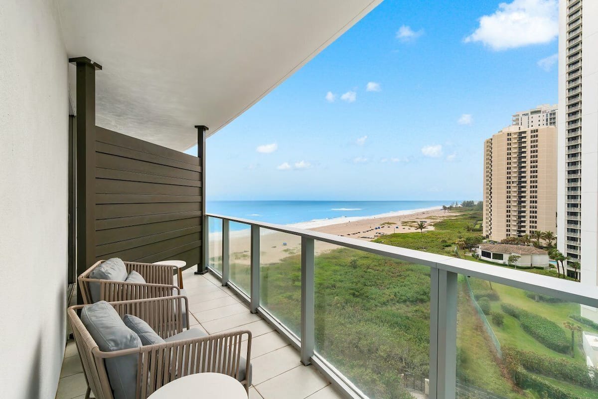 A balcony with wicker furniture, a glass railing, and a view of the beach and ocean.