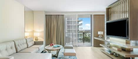 Una habitación de hotel moderna con sofá, televisor, balcón con vistas a edificios y un ventanal grande con cortinas.