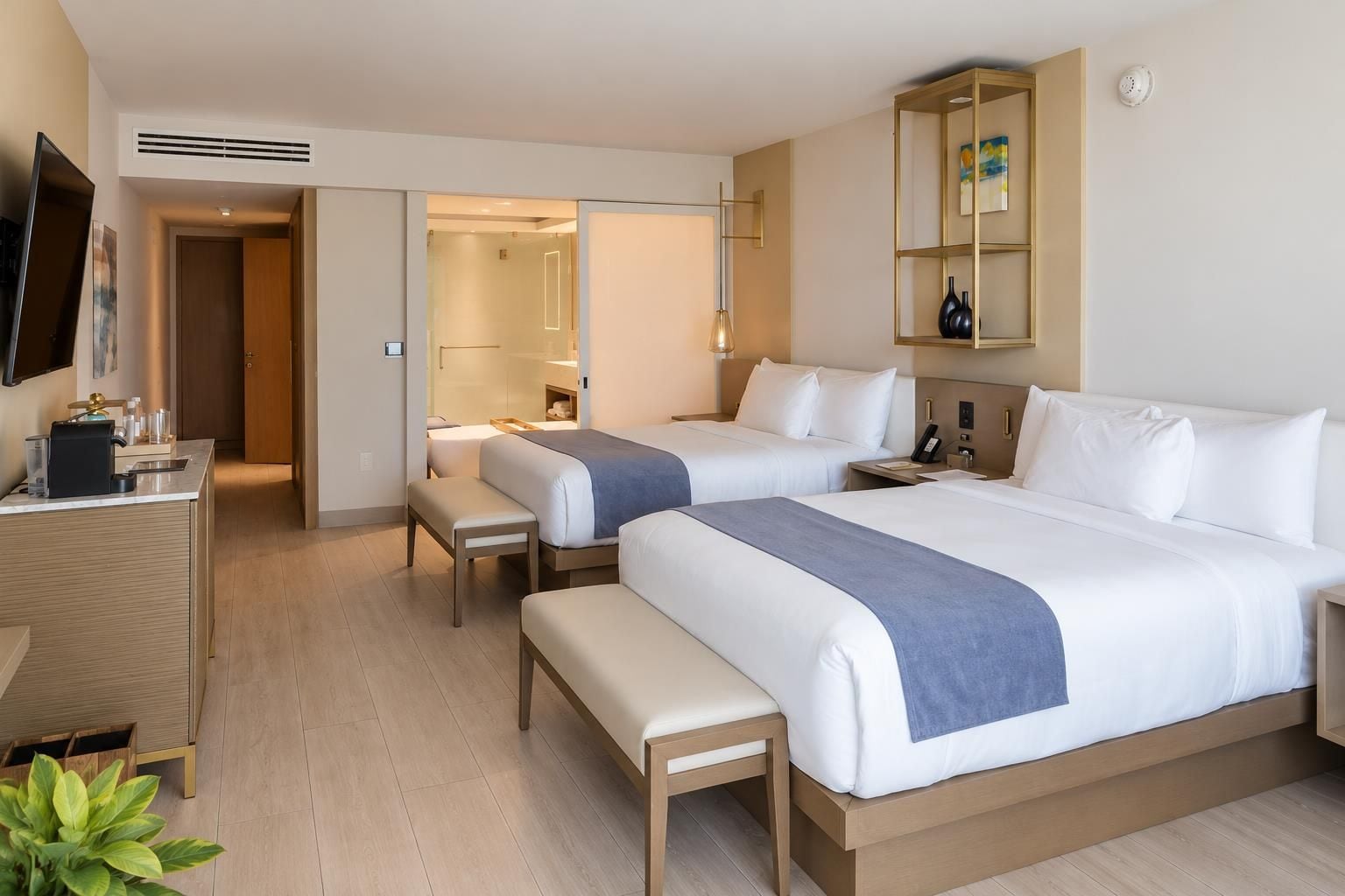 A hotel room with two beds, a desk, a bench, a TV, and a mirror.