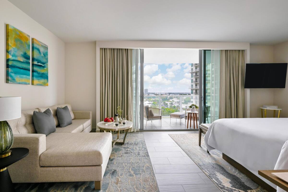 A hotel room with a sofa, a bed, a TV, a balcony with a view of the city, and artwork on the wall.