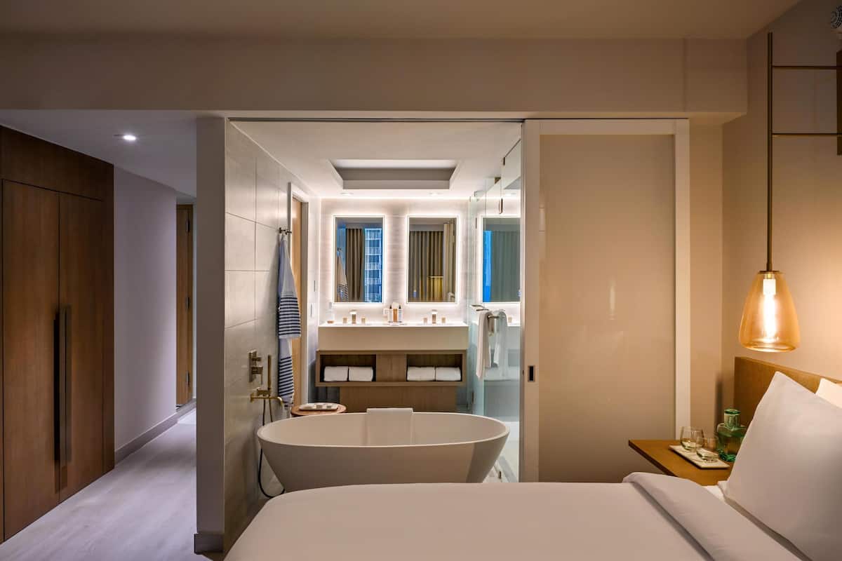 A modern hotel room with a large bed, a bathtub, a vanity with a mirror, and a small table with a bottle and glass.