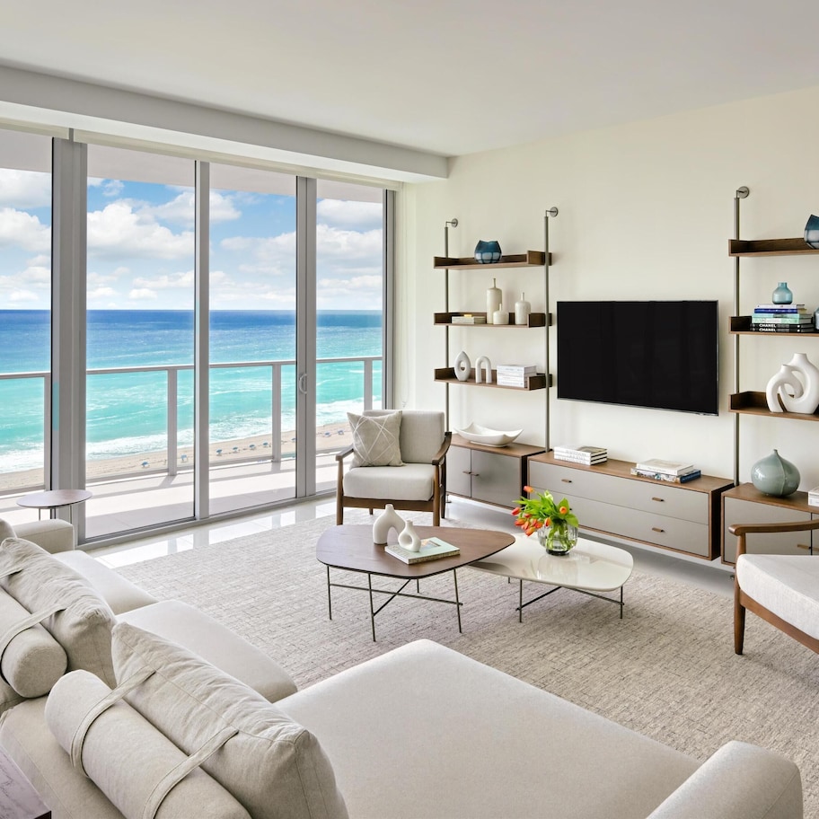 A spacious living room with a large window overlooking the ocean, a comfortable sofa, a coffee table, and a TV mounted on the wall.