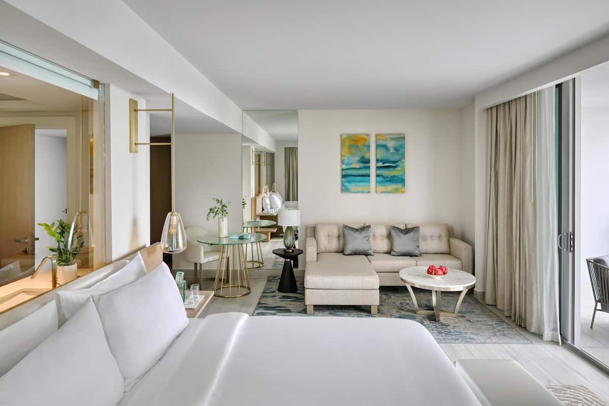 A modern hotel room with a large bed, a sofa, a coffee table, and a dining area. There is a large mirror, a painting on the wall, and a view of the outside.