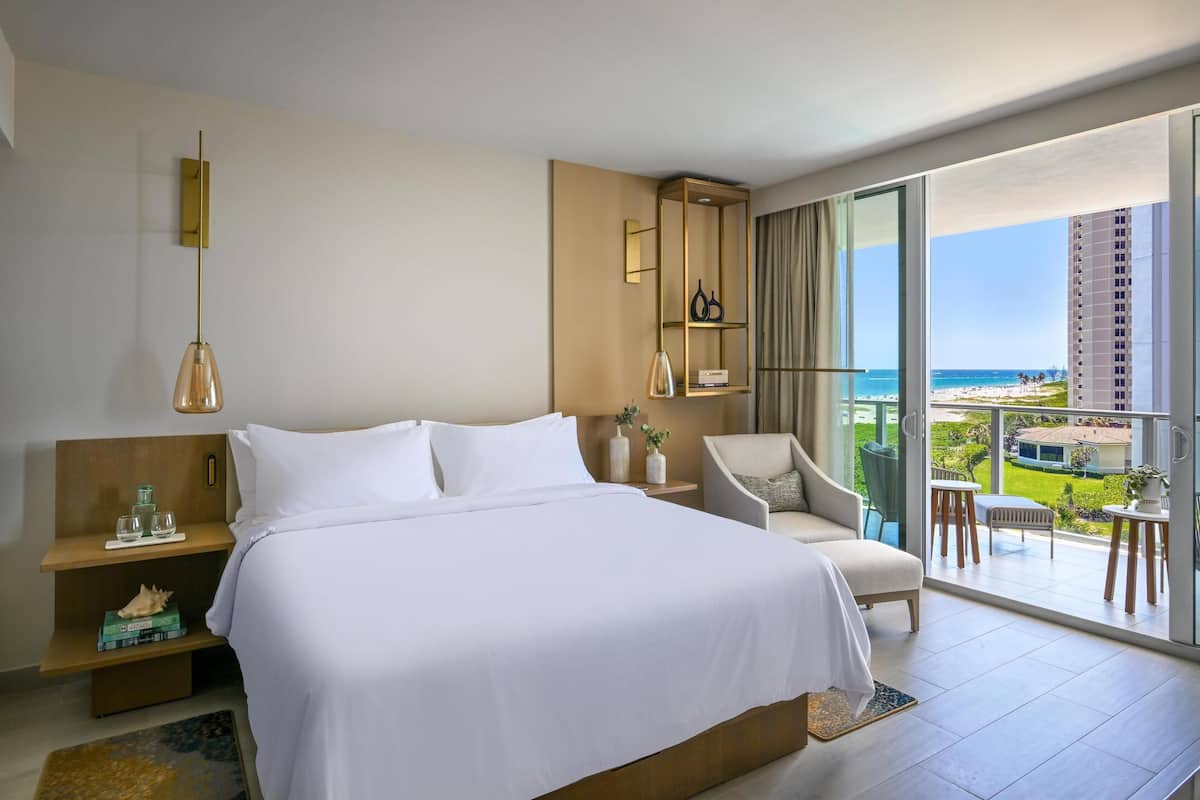 A hotel room with a large bed, a sitting area with a chair and table, a nightstand, a lamp, and a view of the beach and ocean through large windows.