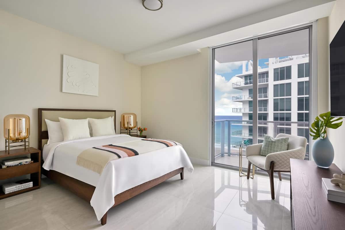 A modern hotel room with a large bed, a bedside table with a lamp, a chair, a view of a building through a large window, and a potted plant.