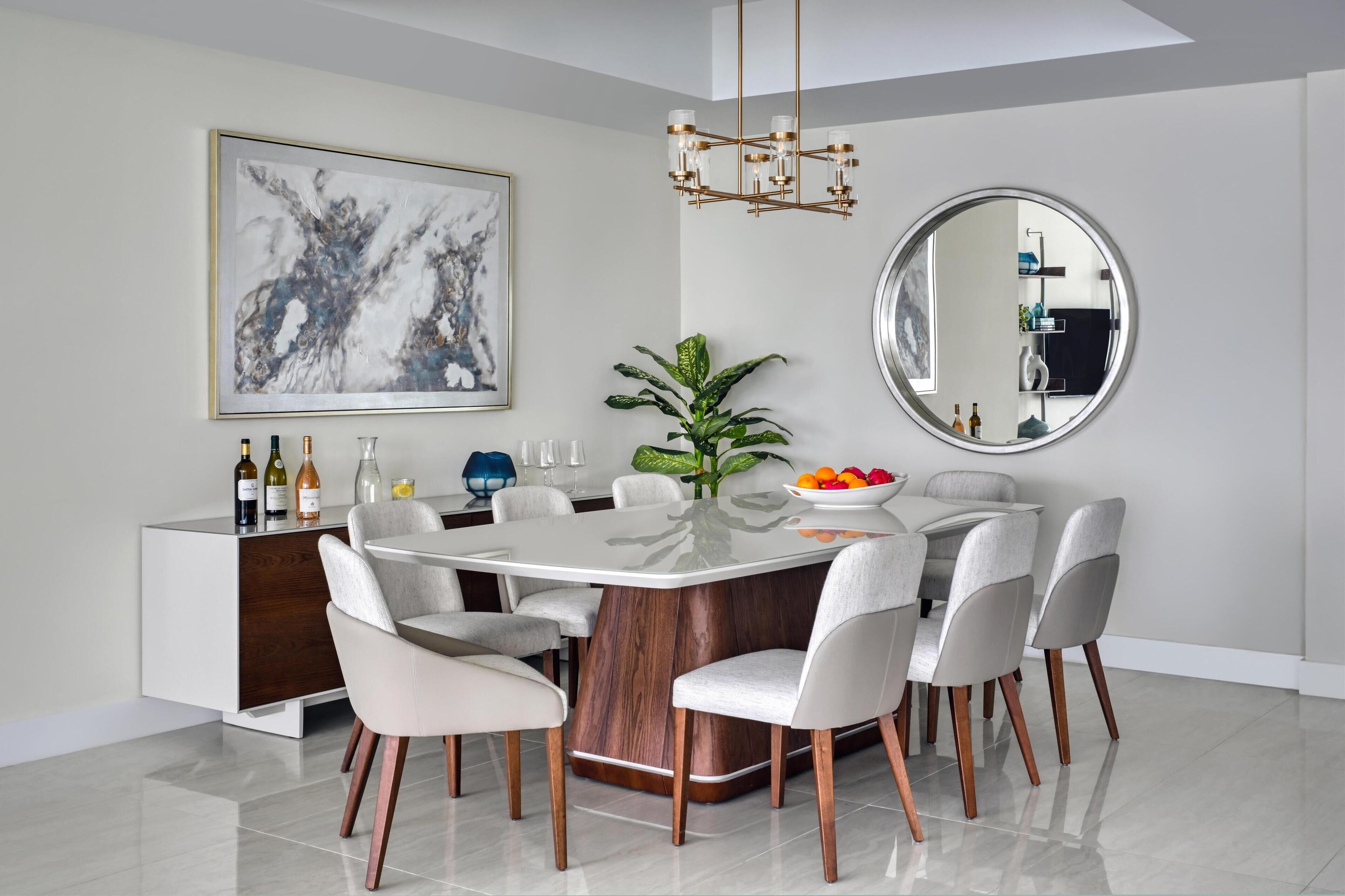 A modern dining room with a round table, white chairs, a wooden cabinet, a large abstract painting, a mirror, and a potted plant.