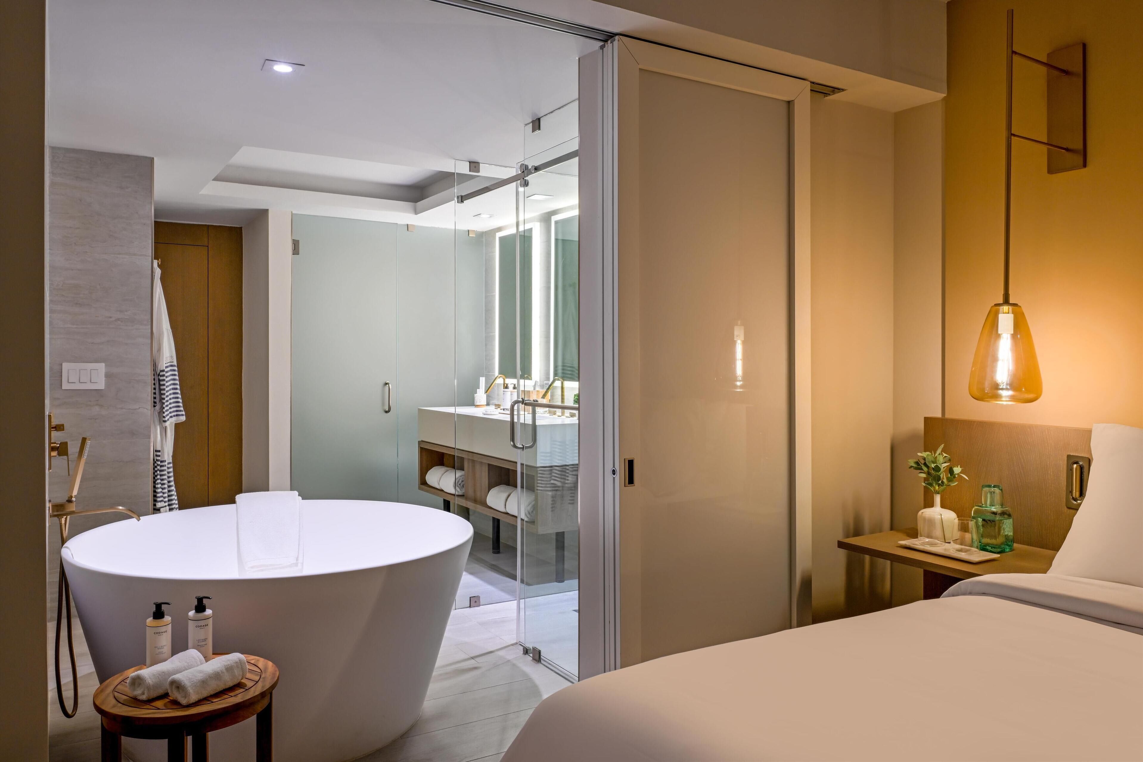 A modern hotel room with a large round bathtub, a separate shower area, a bedside table with a lamp, and a bed with a headboard.