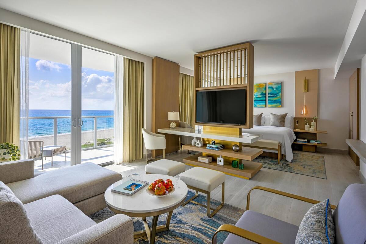 A modern hotel room with a balcony, a bed, a TV, and a dining area.