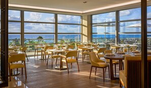A spacious dining area with large windows overlooking the ocean, wooden flooring, and yellow chairs.