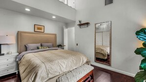 A bedroom with a bed, bedside table, lamp, mirror, and a large plant.