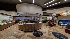 Un hall d’hôtel moderne avec un comptoir d’accueil central, un espace salon et un restaurant en arrière-plan.