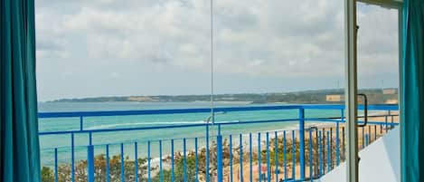 A room with a large window overlooking a beach and ocean, featuring a balcony with blue railings.
