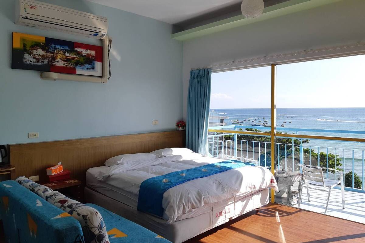 A hotel room with a bed, a small table, a chair, and a balcony with a view of the sea.
