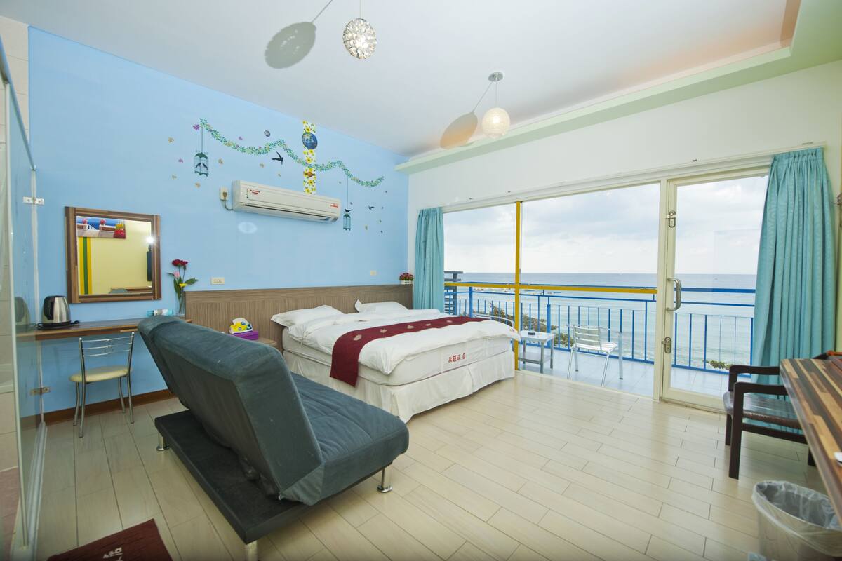A hotel room with a bed, a desk, a chair, a balcony with a view of the sea, and a window with curtains.