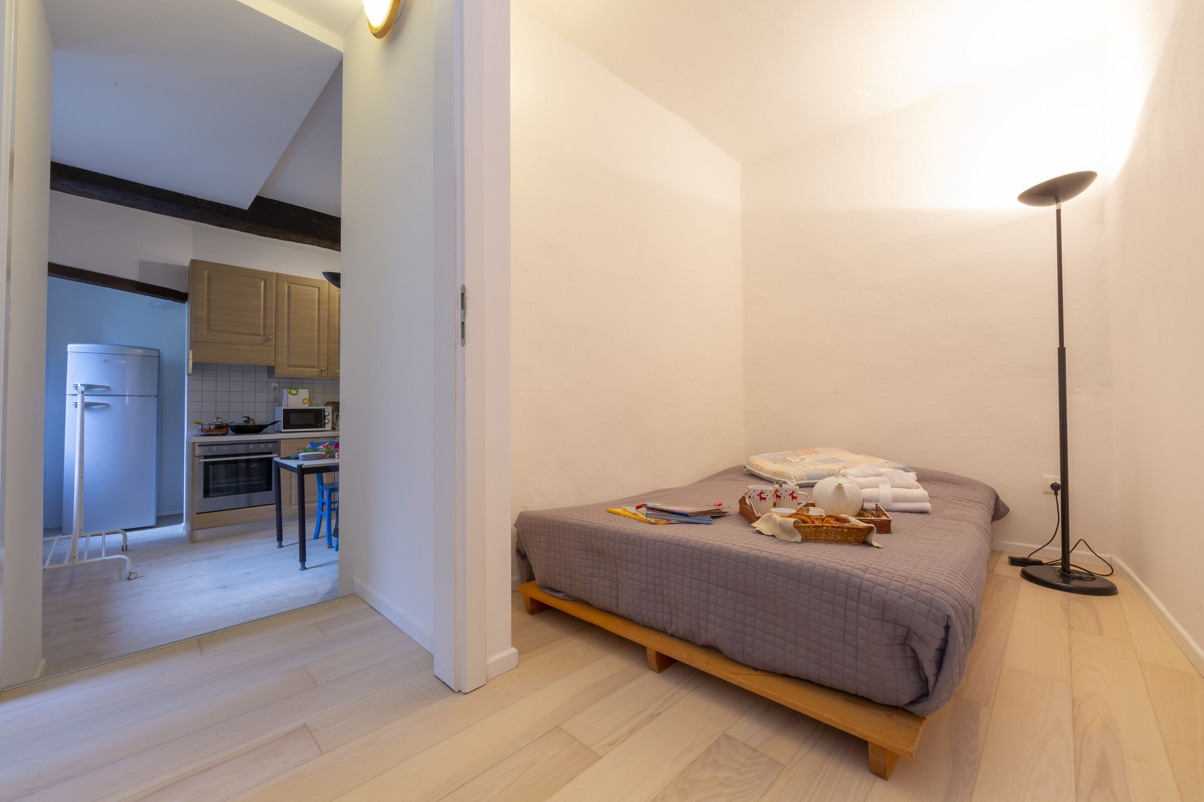 Un dormitorio con cama, mesita de noche y lámpara de pie. Junto al dormitorio, hay una cocina con armarios y electrodomésticos.
