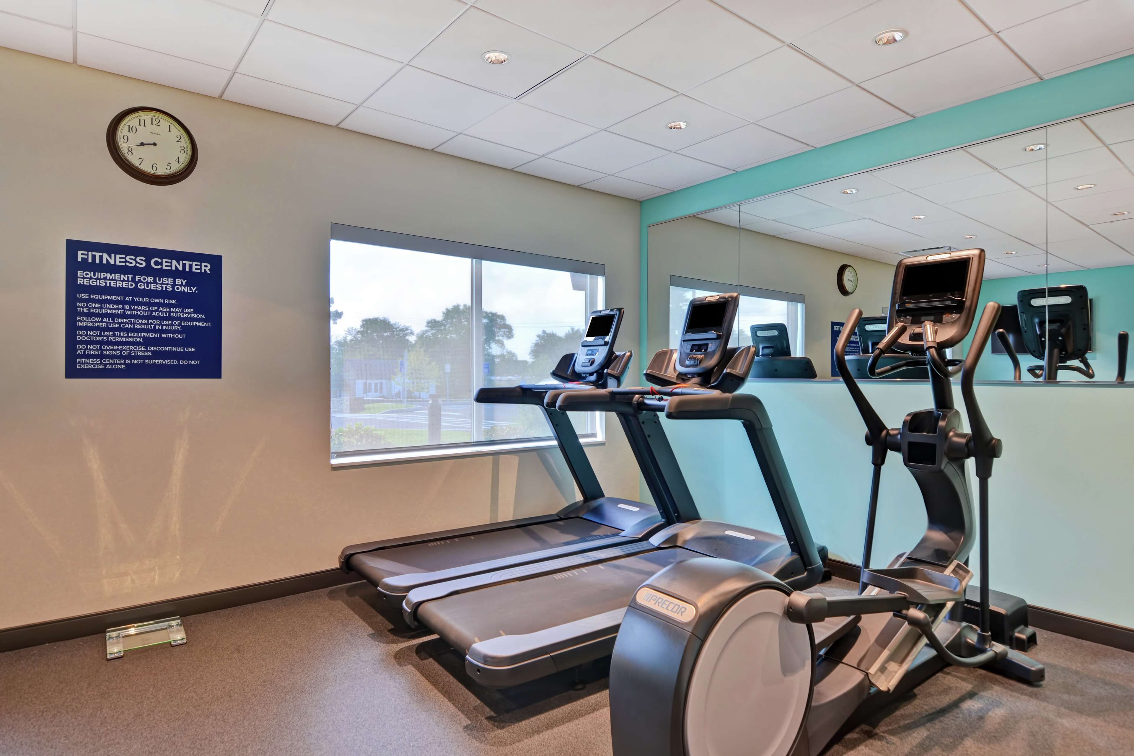 A fitness center with treadmills, an elliptical, and a stationary bike. There's a large window, a clock, and a sign with equipment usage instructions.