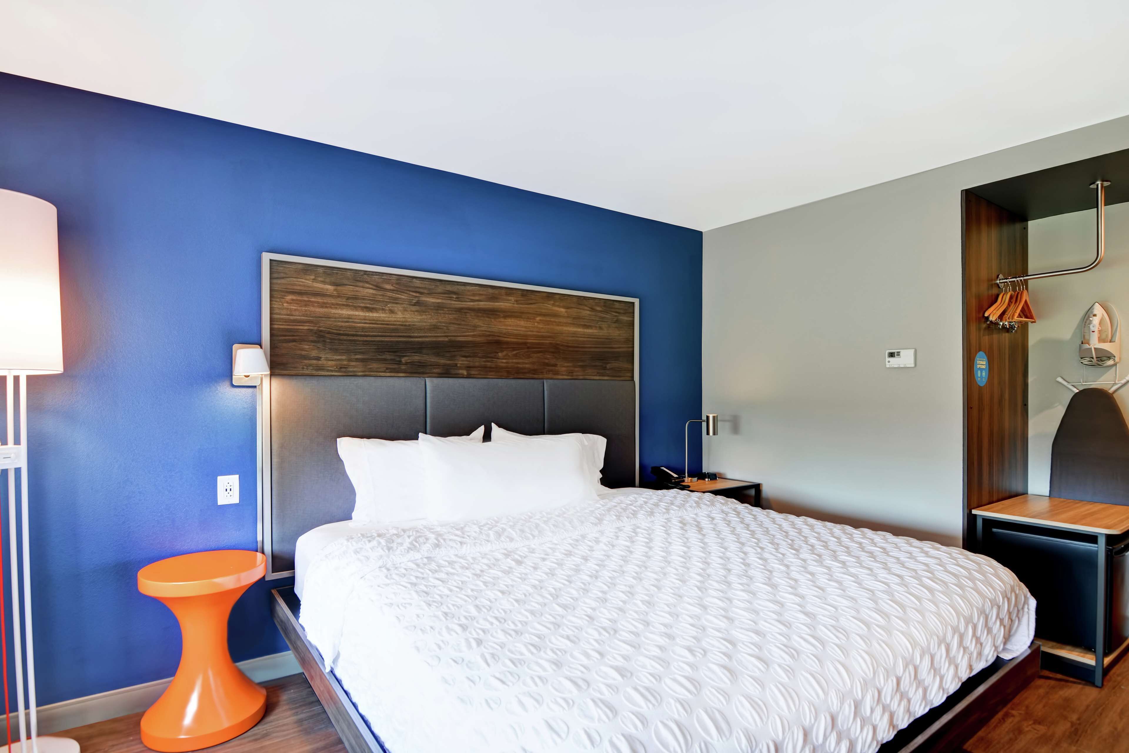Una habitación de hotel moderna con una cama grande, un cabecero de madera, una mesita de noche naranja pequeña y un espejo con toallero.