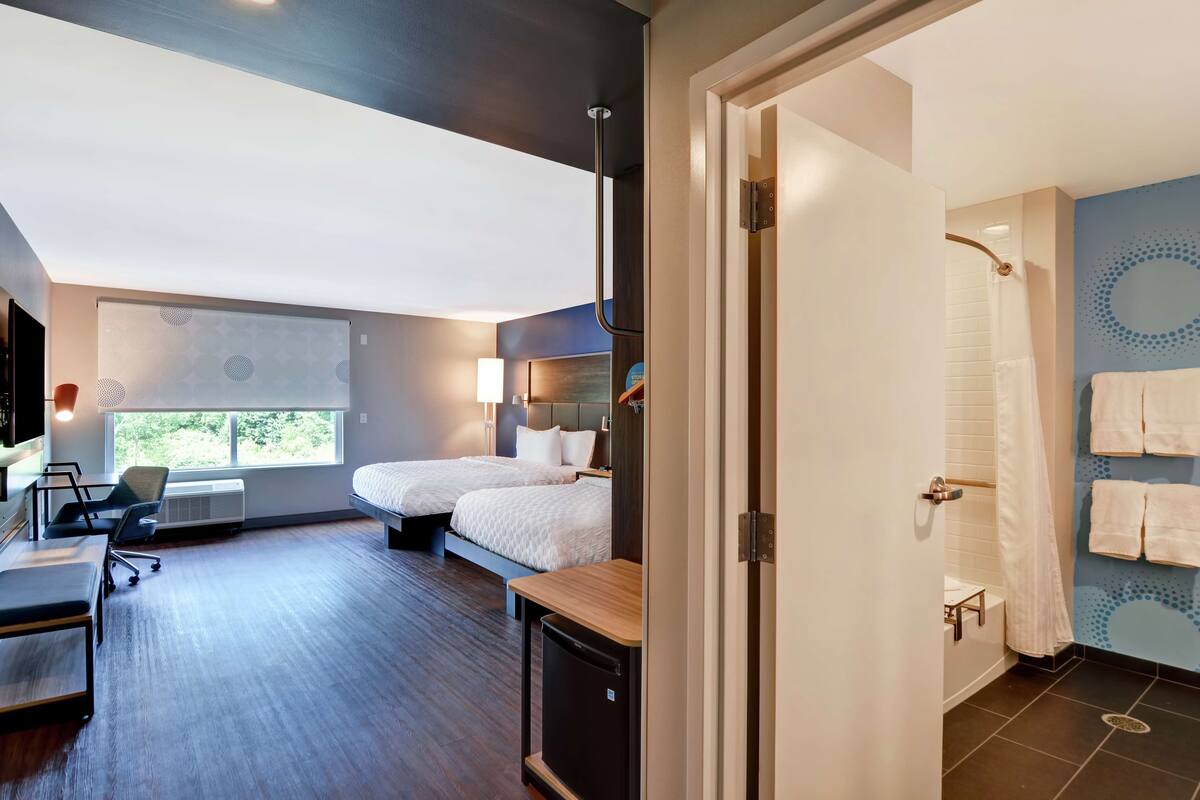 A modern hotel room with two beds, a desk, a chair, and a view of the outdoors.