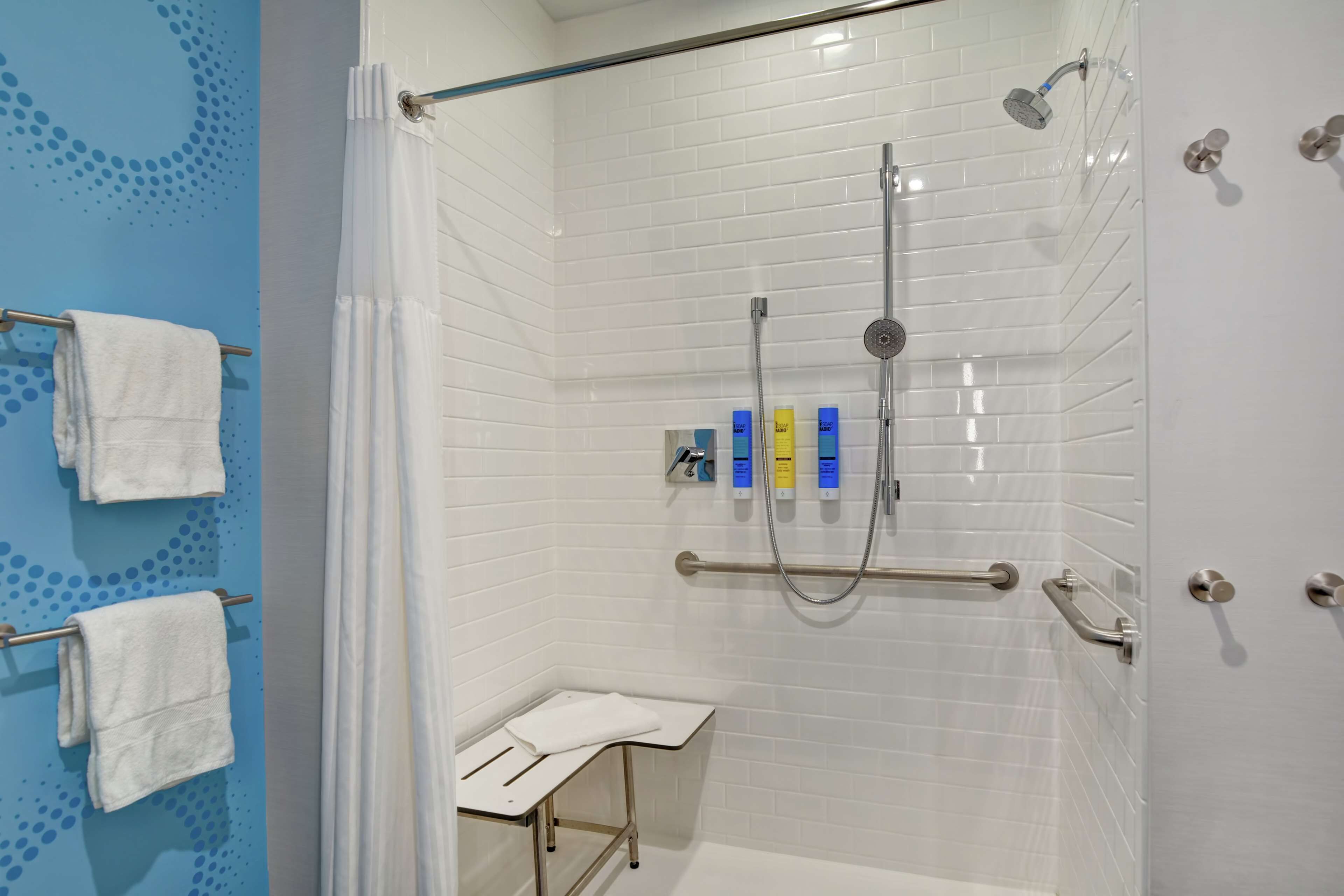 Zona de ducha con paredes de azulejos blancos, puerta de vidrio y cabezal de ducha. Hay tres botellas de champú y acondicionador en la pared.