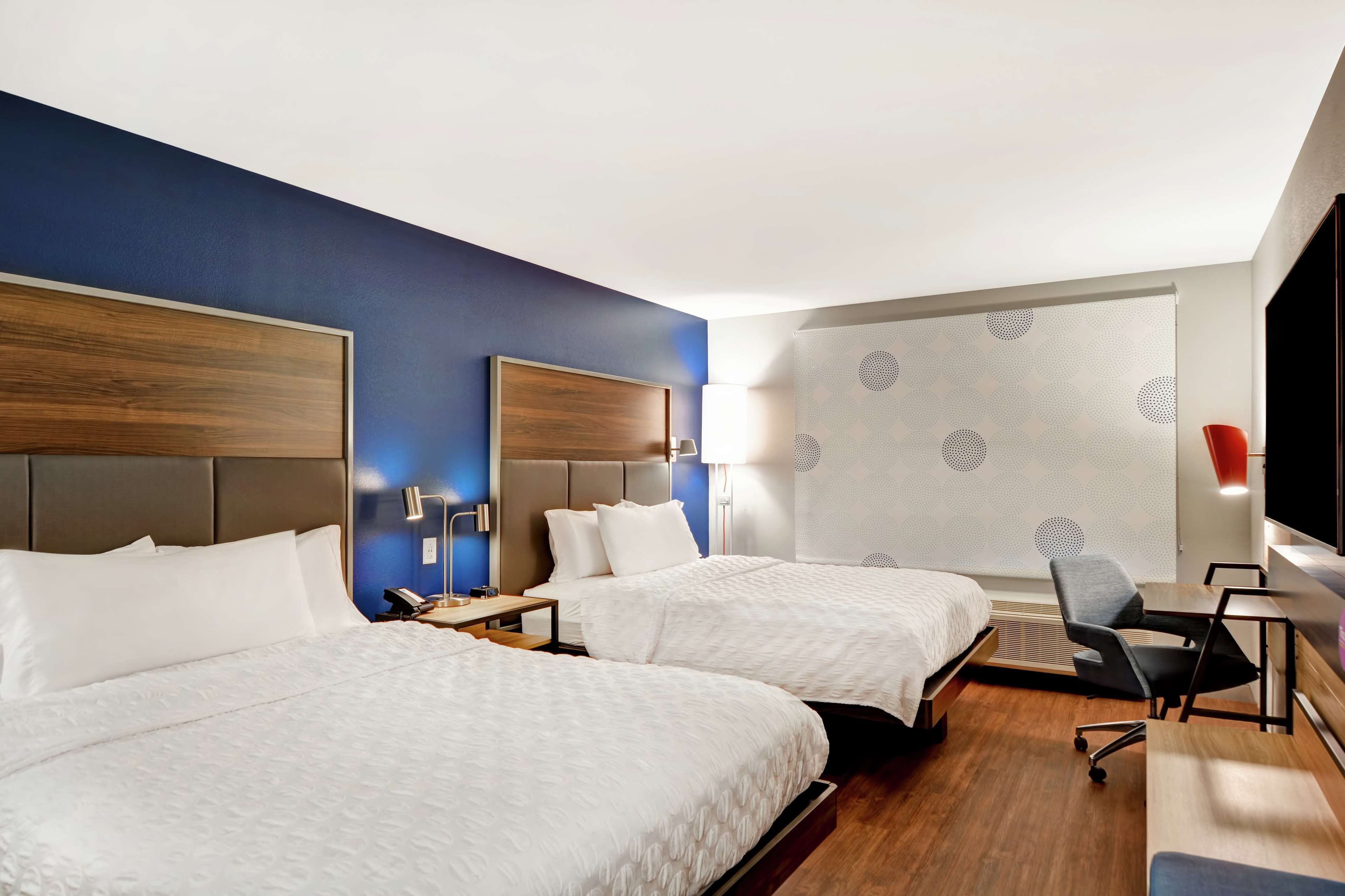 A hotel room with two beds, a desk, a chair, and a large window with a patterned curtain.