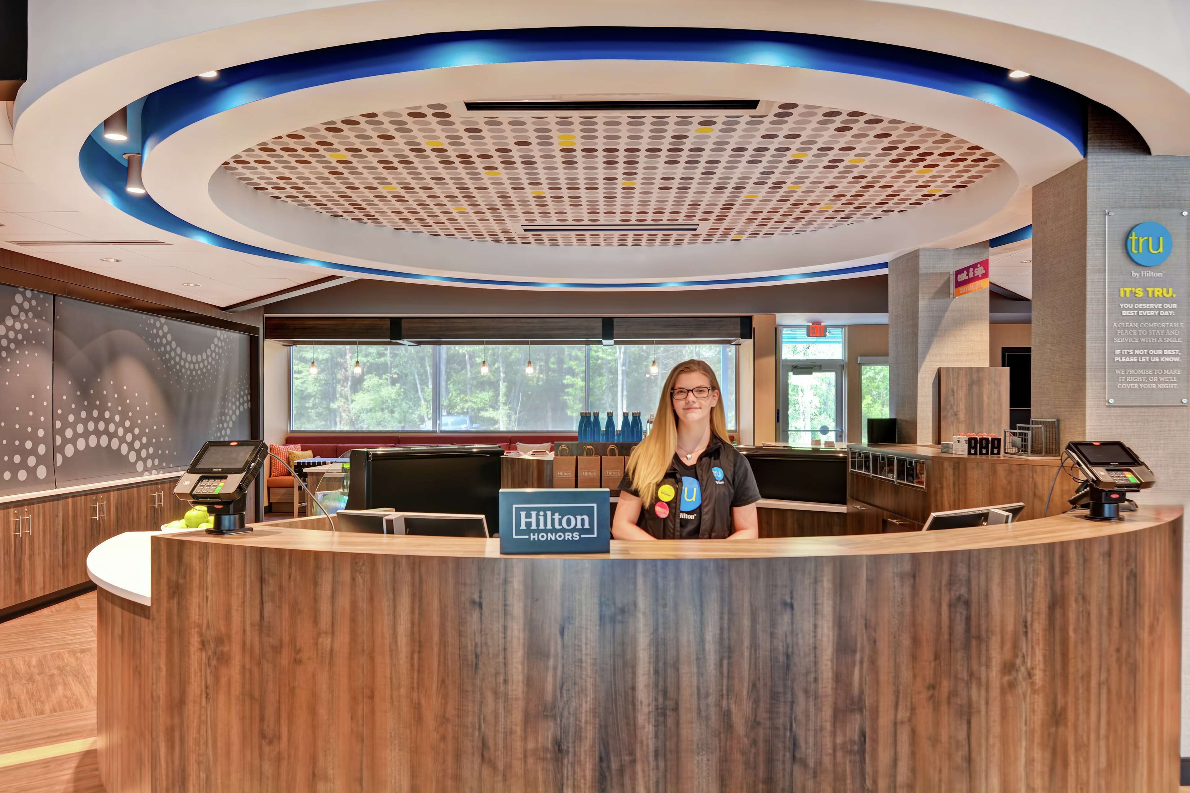 A modern hotel reception desk with a digital kiosk, a sign indicating Hilton Honors, and a view of the outdoors through large windows.